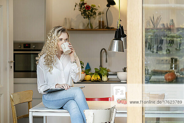 Young freelancer drinking coffee sitting with laptop on table in kitchen