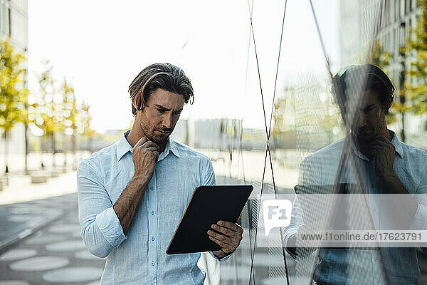 Businessman with hand on chin using tablet PC by office building