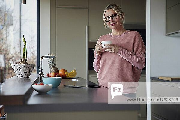 Blonde Frau mit Brille und Kaffeetasse steht in der Küche