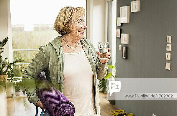Happy senior woman with exercise mat holding glass of water at home