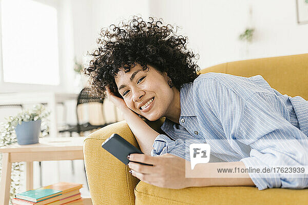 Happy woman holding mobile phone lying on sofa in living room