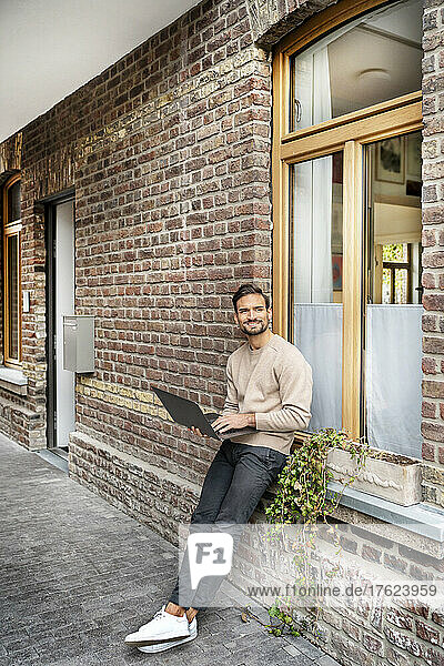 Smiling freelancer with laptop leaning on window sill