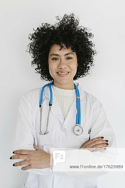 Happy young doctor standing with arms crossed in front of white wall