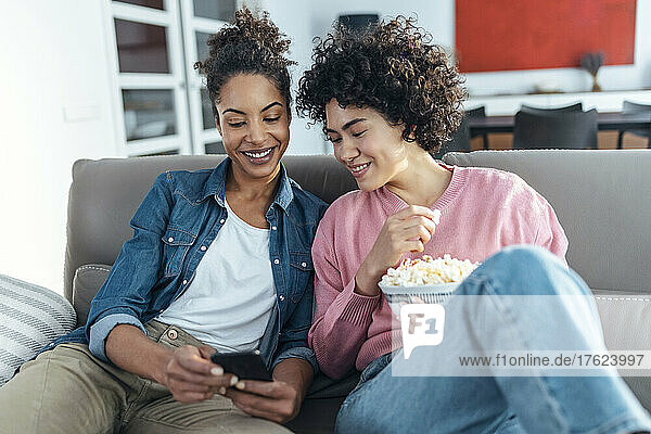 Smiling woman showing smart phone to friend sitting on sofa at home