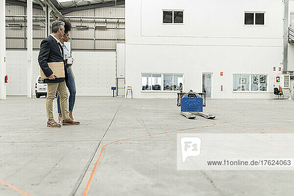 Businessman holding laptop standing with businesswoman looking at pallet jack in factory