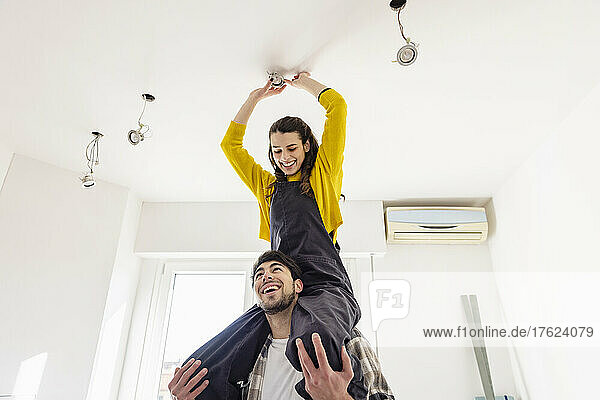 Boyfriend carrying girlfriend on shoulders installing light bulb at new home