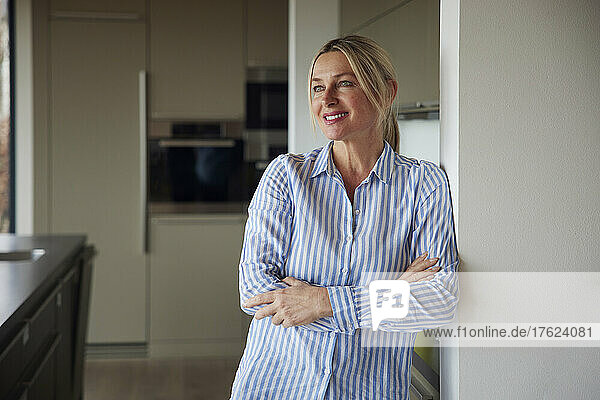 Happy blond woman standing with arms crossed leaning on wall at home