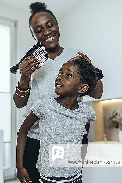 Smiling mother combing daughter's hair in bathroom at home
