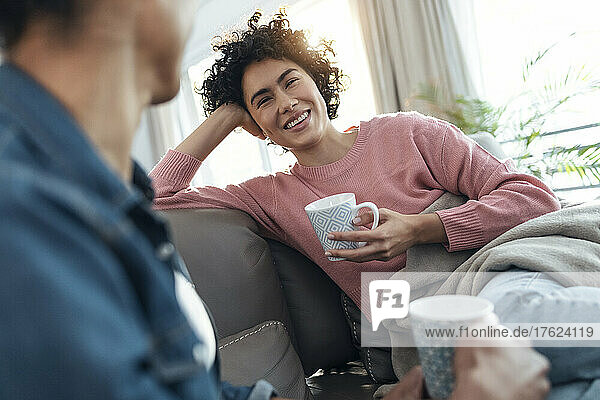 Happy young woman holding coffee cup looking at friend sitting on sofa at home