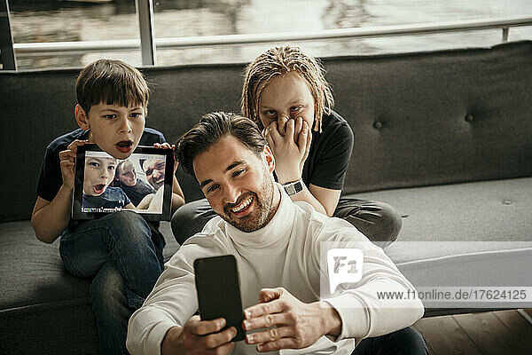Happy man taking selfie through smart phone with sons making faces at home