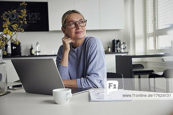 Nachdenkliche Geschäftsfrau mit Brille sitzt mit Laptop am Tisch