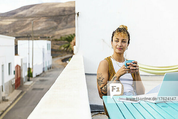 Young woman holding tea cup sitting with laptop on rooftop