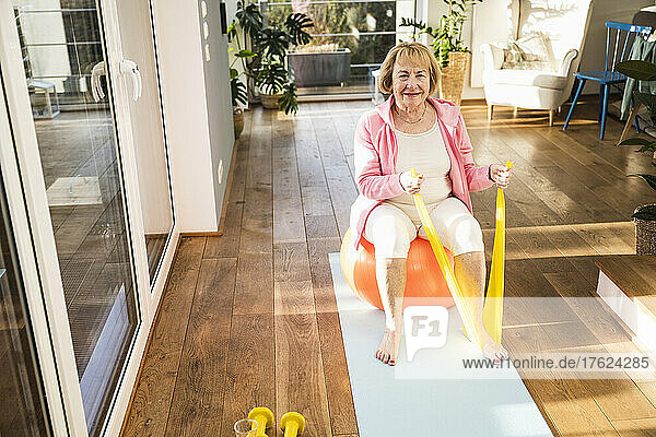 Smiling senior woman sitting on fitness ball exercise with resistance band at home