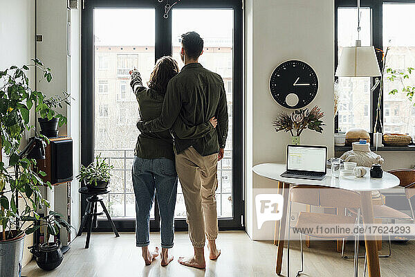 Young couple standingwith arms around by glass window at home