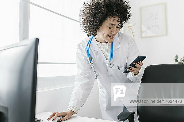 Smiling doctor with curly hair using smart phone at clinic