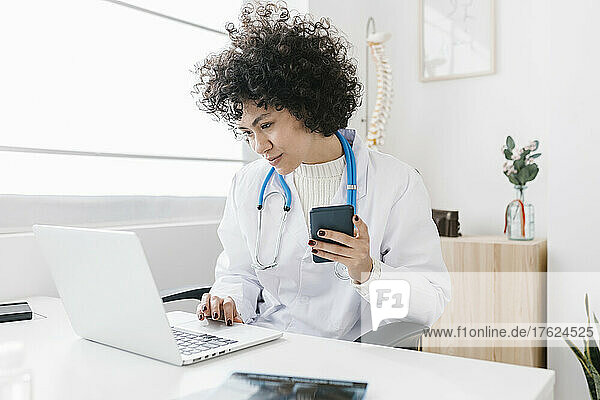 Young doctor holding smart phone using laptop at clinic