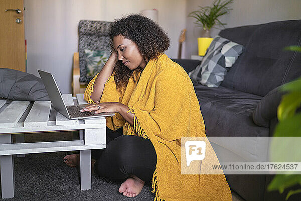 Young woman wrapped in yellow blanket using laptop sitting at coffee table at home