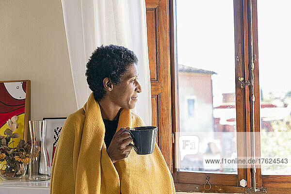 Thoughtful woman with blanket having coffee by window at home