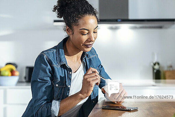 Woman eating yogurt sitting in kitchen at home
