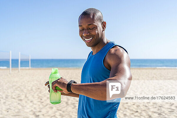 Smiling man holding water bottle checking time at beach on sunny day