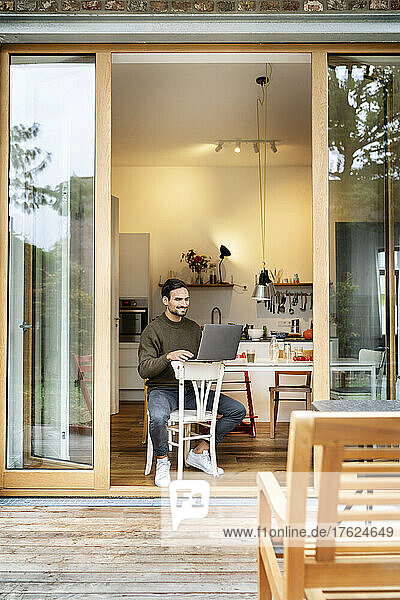 Smiling freelancer using laptop sitting on chair working in kitchen