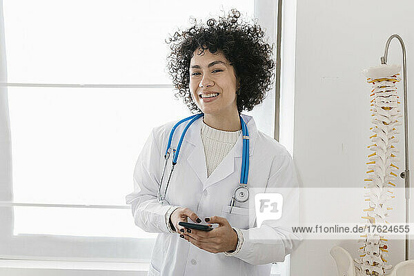 Happy young doctor holding smart phone standing by window at clinic