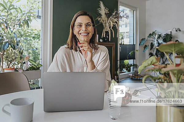 Smiling freelancer with strawberry sitting with laptop at desk in home office