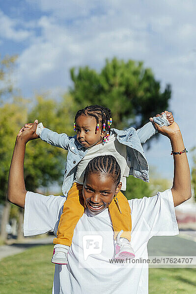 Smiling young woman carrying cute daughter on shoulders at park