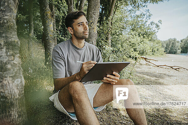 Thoughtful man with tablet PC sitting at tree