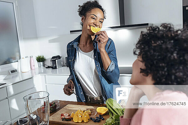 Smiling woman covering mouth with slice of orange looking at friend sitting in kitchen