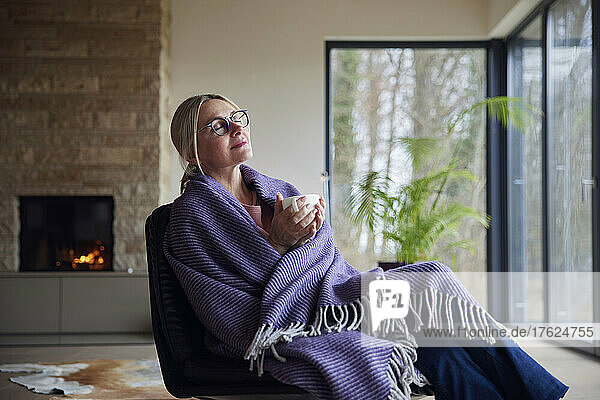 Entspannte Frau  in eine Decke gehüllt  sitzt mit geschlossenen Augen im Wohnzimmer