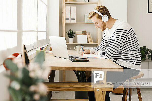 Young man listening music through wireless headphones writing in book siting at table