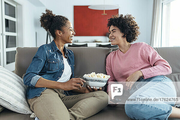 Happy woman holding bowl of popcorn talking with friend on sofa at home