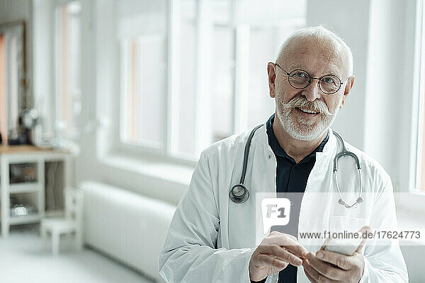 Smiling senior doctor holding mobile phone at hospital