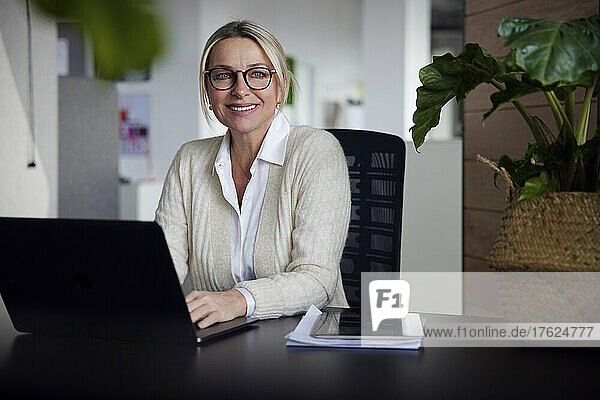 Smiling businesswoman with laptop sitting at desk in office