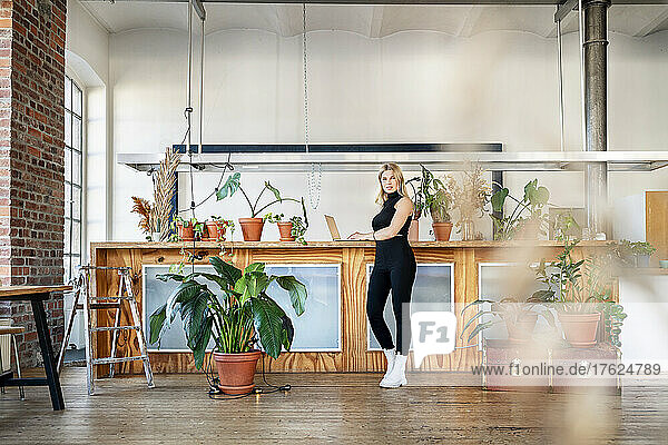 Businesswoman with laptop standing by potted plants in living room at home