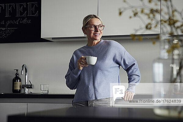 Smiling woman holding coffee cup leaning on kitchen counter