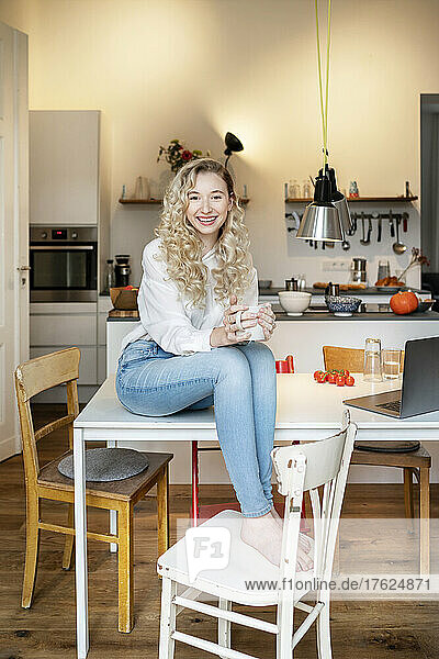 Glückliche schöne Frau mit blonden Haaren hält eine Kaffeetasse in der Hand und sitzt auf dem Tisch in der Küche