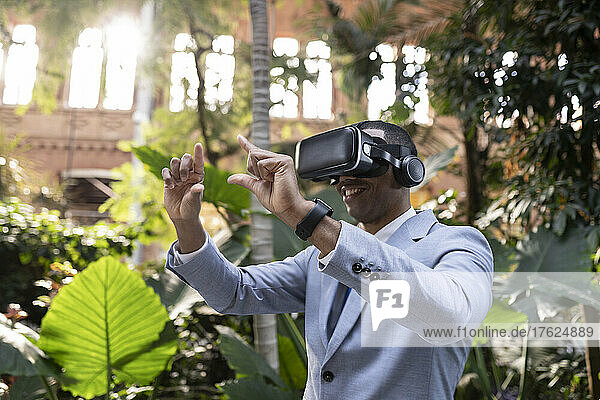 Smiling businessman wearing virtual reality simulator gesturing hands in front of plants