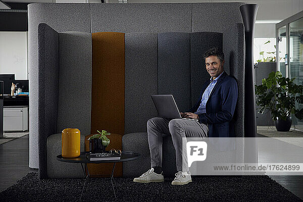 Smiling businessman with laptop sitting on sofa in office