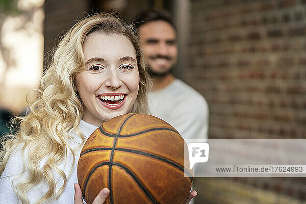 Cheerful blond woman holding basketball in front of boyfriend