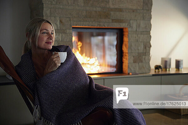 Woman wrapped in blanket holding coffee cup sitting in living room