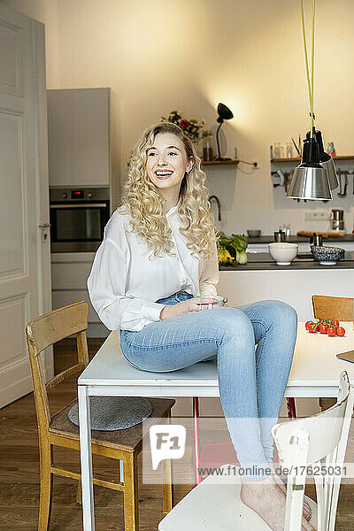 Cheerful blond woman holding coffee cup sitting on table in kitchen at home