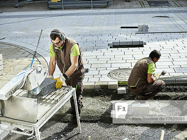 Paver cutting paving stone by coworker working in background