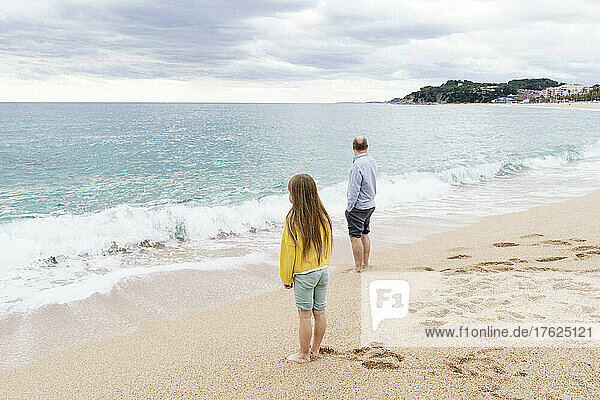 Father and daughter standing by coastline at beach