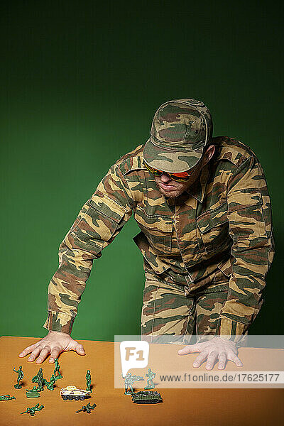 Young military soldier with army figurine toys standing by table against green background
