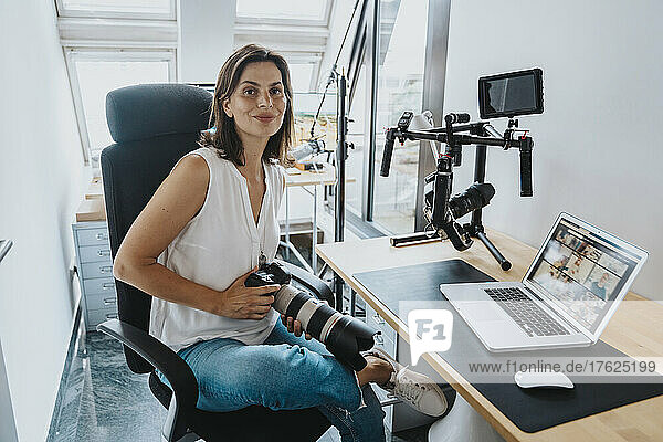 Lächelnder Fotograf mit Digitalkamera sitzt auf einem Stuhl mit Laptop auf dem Tisch im Studio