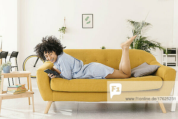 Happy young woman surfing net through smart phone lying on sofa in living room at home