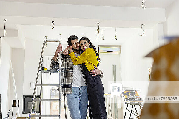 Glückliche Frau umarmt ihren Freund  der an der Leiter im neuen Wohnzimmer steht