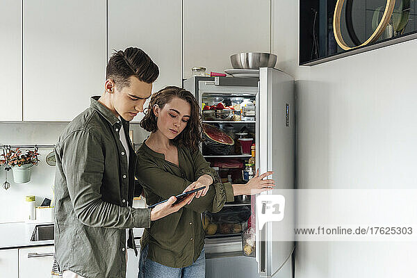 Young man with woman checking list on tablet PC by refrigerator in kitchen